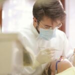 Dentiste portant un masque facial effectuant un examen dentaire avec des outils en milieu clinique.