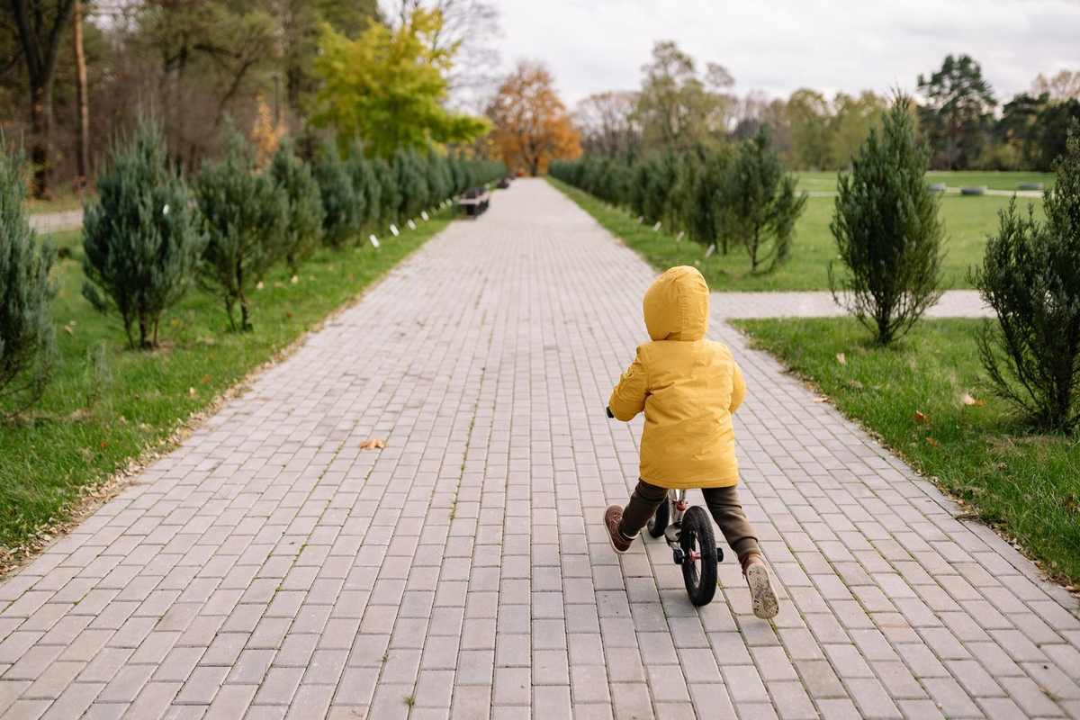 Enfant en manteau jaune sur vélo, chemin pavé, allées d'arbres, ciel gris, automne