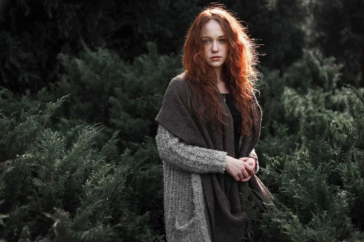 Jeune femme aux cheveux rouges dans une forêt, vêtue d'un cardigan gris et d'un manteau brun, les mains jointes